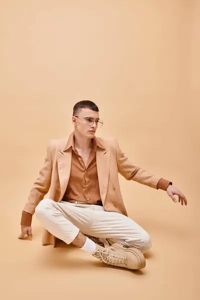 Hombre con estilo en chaqueta beige y gafas sentadas en pose de loto sobre fondo beige melocotón - foto de stock