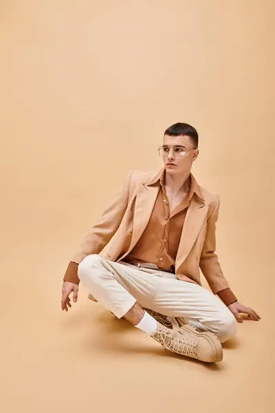 Foto de moda de hombre en chaqueta beige y gafas sentadas en pose de loto sobre fondo beige melocotón - foto de stock