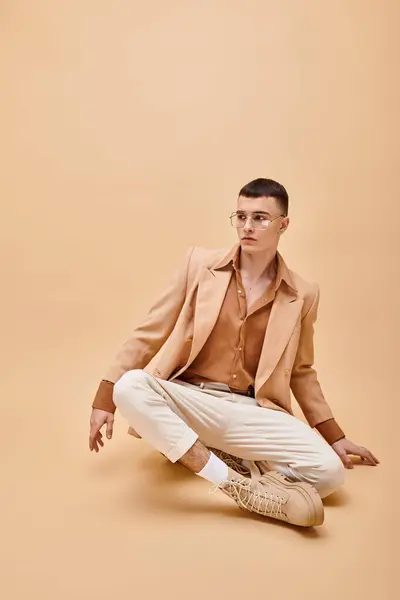 Foto de moda de hombre en chaqueta beige y gafas sentadas en pose de loto sobre fondo beige melocotón - foto de stock