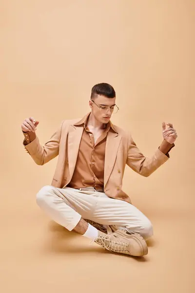 Модный портрет человека в бежевой куртке и очках, сидящих на персиковом бежевом фоне — стоковое фото