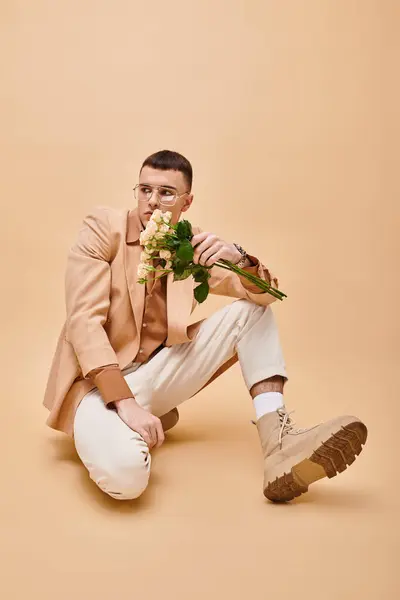 Foto de moda de hombre en chaqueta beige y gafas sentadas con flores sobre fondo beige turquesa - foto de stock