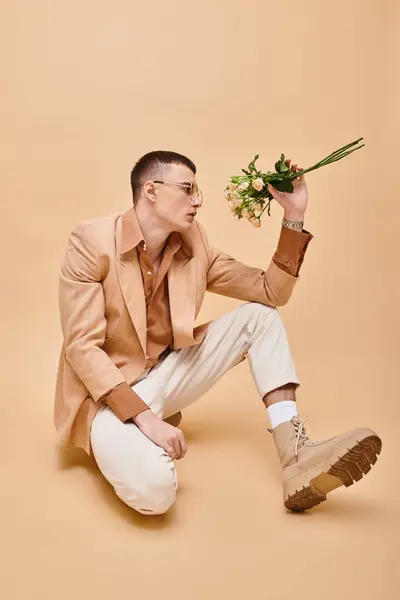 Fashion portrait of man in beige jacket and glasses sitting with roses on peachy beige background - foto de stock