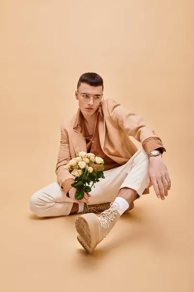 Bello uomo in giacca beige e occhiali seduto con rose su sfondo beige pesca — Foto stock