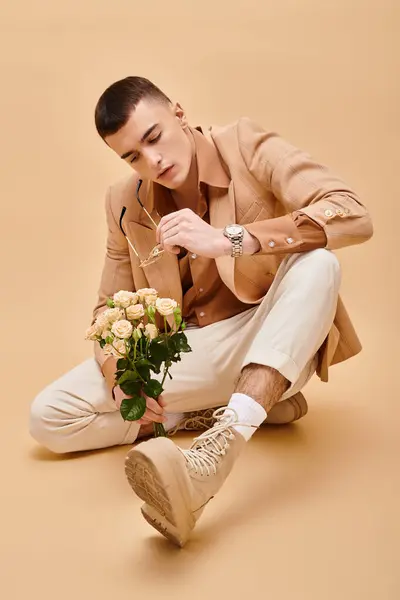 Fashion shot of man in beige jacket sitting with roses and eyeglasses on peachy beige background — Stock Photo