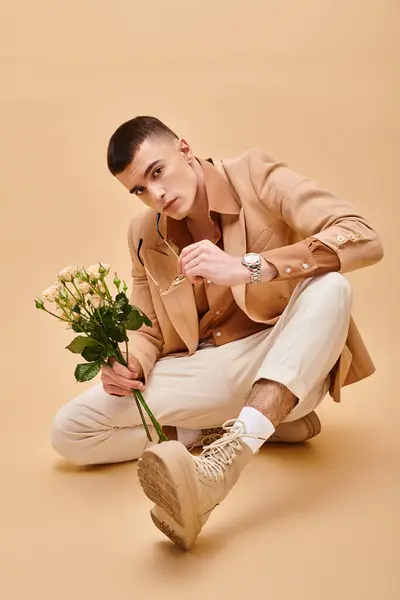 Schöner Mann in beiger Jacke sitzt mit Rosen auf pfirsichfarbenem beigem Hintergrund und blickt in die Kamera — Stockfoto