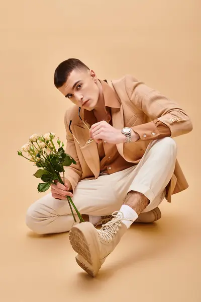 Beau homme en veste beige assis avec des roses sur fond beige pêche regardant la caméra — Photo de stock