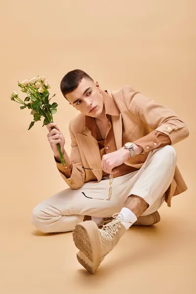 Hombre con estilo en chaqueta beige sentado con rosas y gafas sobre fondo beige mirando a la cámara - foto de stock