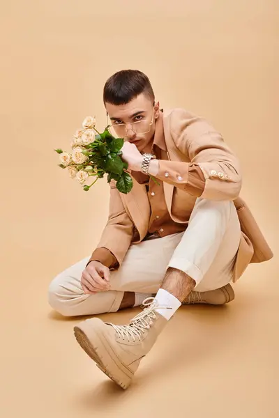 Young man in beige jacket sitting with roses and glasses on beige background looking at camera — Stock Photo