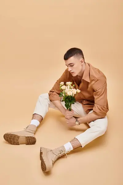 Hombre de moda en camisa beige sentado con flores y gafas sobre fondo beige mirando hacia abajo - foto de stock