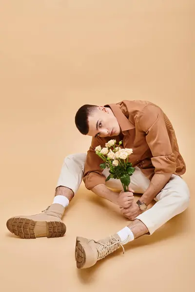 Jeune homme en chemise beige assis avec des fleurs et des lunettes sur fond beige regardant la caméra — Photo de stock