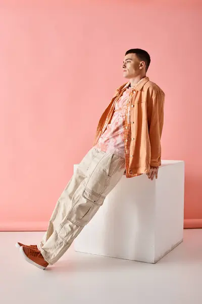 Fashion shot of fashionable man in beige shirt, pants and boots on white cube on pink background — Stock Photo
