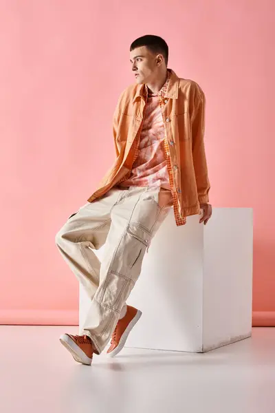 Fashionable man in beige shirt, pants and boots sitting on white cube on pink background — Stock Photo
