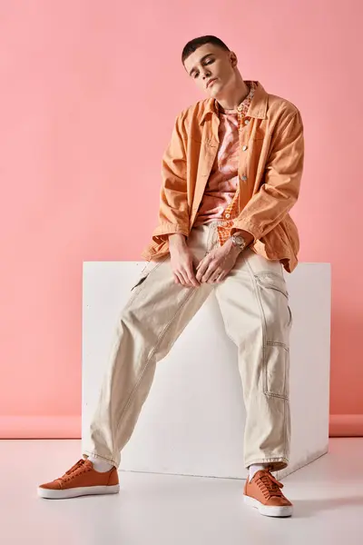Handsome man in beige shirt, pants and sneakers sitting on white cube on pink background — Stock Photo
