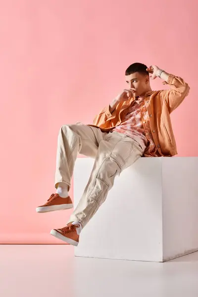 Young man in beige outfit looking away and sitting on white cube on pink background — Stock Photo