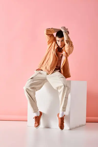 Uomo alla moda con le mani sopra la testa e seduto su cubo bianco su sfondo rosa — Foto stock