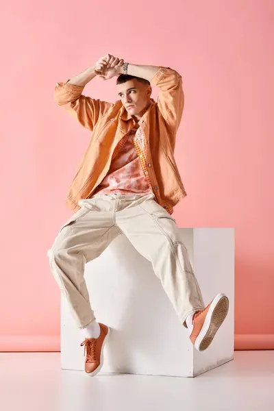 Fashionable young man in beige outfit touching his head and sitting on white cube on pink background — Stock Photo