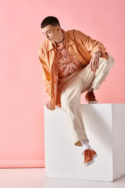 Stylish man in beige outfit looking down and sitting on white cube on pink background — Stock Photo