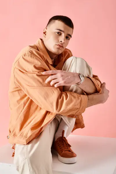 Hombre de 20 años en traje elegante abrazando escondido rodilla y sentado en cubo blanco sobre fondo rosa - foto de stock