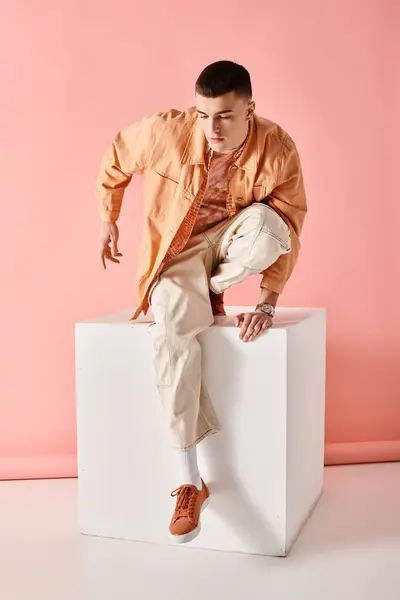 Hombre guapo en traje beige mirando hacia abajo y posando sobre cubo blanco sobre fondo rosa - foto de stock