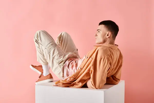 Stylish man in peachy outfit touching his hair and lying on white cube on pink background — Stock Photo