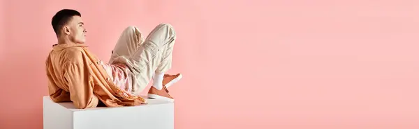 Stylish man in peachy outfit touching his hair and lying on white cube on pink background, banner — Stock Photo