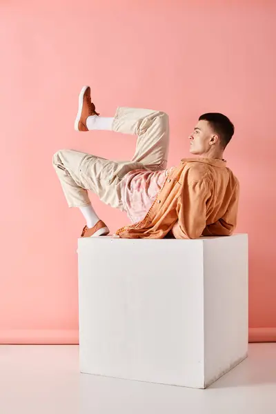 Seitenansicht Foto von Mann in pfirsichfarbenem Outfit auf weißem Würfel liegend mit Bein nach oben auf rosa Hintergrund — Stockfoto