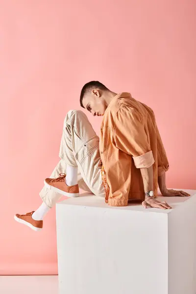 Side view portrait of man in peach color outfit lying on white cube with knee up on pink background — Stock Photo