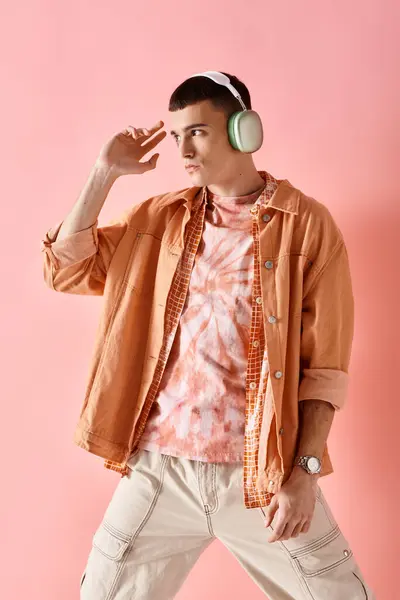 Молодой человек в многослойных рубашках с беспроводными наушниками, слушающий музыку, позирующую на розовом фоне — стоковое фото