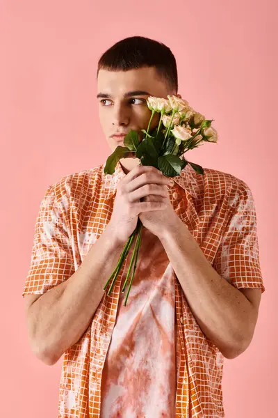 Handsome man in layered peach color outfit holding roses near face on pink backdrop - foto de stock