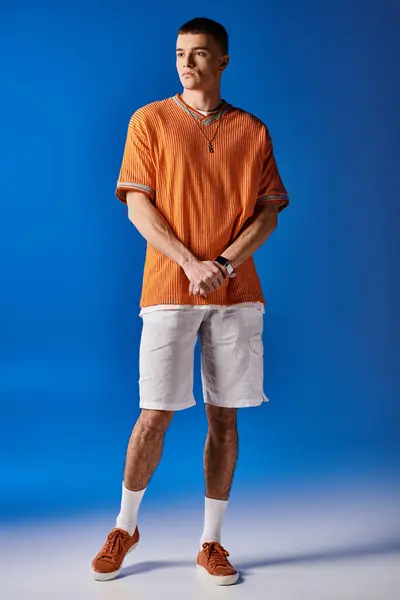 Imagen completa del hombre guapo en camisa naranja y pantalones cortos blancos posando sobre fondo azul - foto de stock