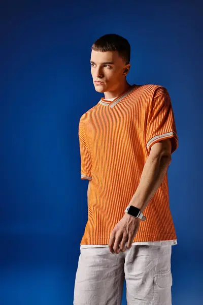 Elegante joven con camisa naranja de moda y pantalones cortos blancos mirando hacia otro lado sobre fondo azul profundo - foto de stock
