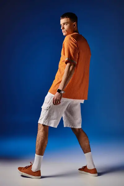 Full length side view shot of handsome man in orange shirt and white shorts posing on blue backdrop — Stock Photo