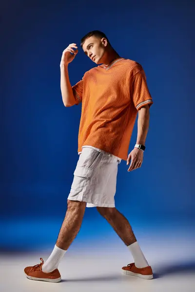 Full length image of handsome man in orange shirt and white shorts posing on blue backdrop — Stock Photo