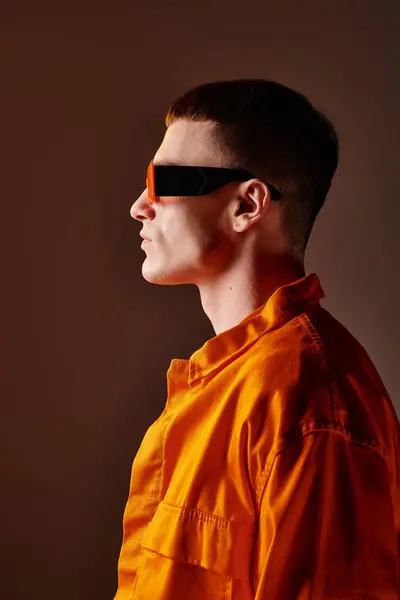 Retrato de moda vista lateral de hombre guapo en camisa naranja y gafas de sol sobre fondo marrón - foto de stock