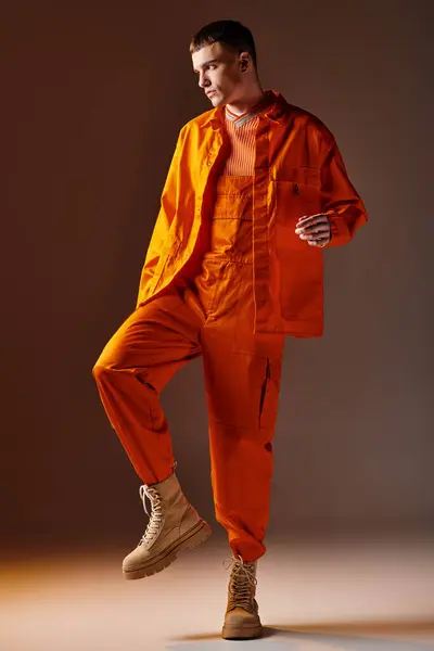Полная длина снимка стильного мужчины в оранжевом комбинезоне и куртке, позирующего на коричневом фоне — стоковое фото