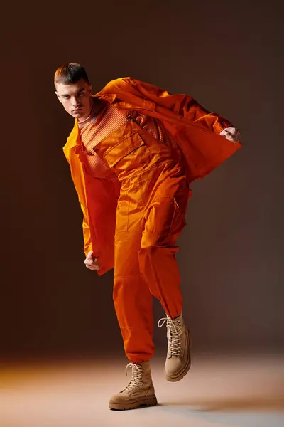 Moda tiro de comprimento total de homem elegante em macacão laranja e casaco posando no fundo marrom — Fotografia de Stock