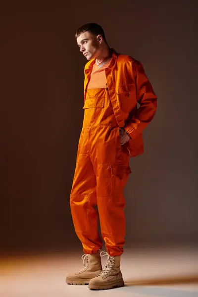 Полнометражный портрет модного человека в оранжевом комбинезоне и куртке, стоящего на коричневом фоне — стоковое фото