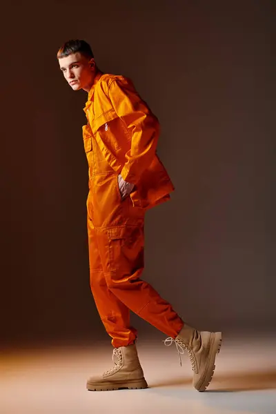 Fashionable guy in 20s in orange jumpsuit and jacket, beige beanie posing on brown background - foto de stock