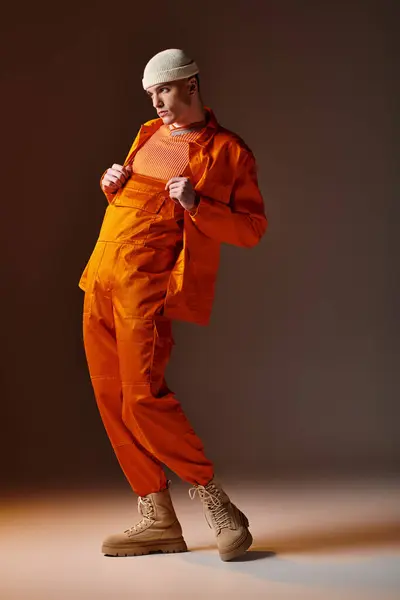 Tiro de comprimento total do homem em macacão laranja e jaqueta, gorro bege posando no fundo marrom — Fotografia de Stock