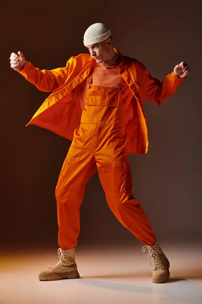 Full length photo of man in orange jumpsuit and jacket, beige beanie dancing on brown background - foto de stock