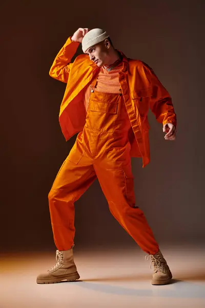 Joven hombre guapo en mono naranja y chaqueta, gorro beige posando sobre fondo marrón - foto de stock
