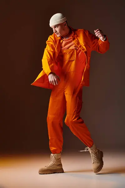 Ganzkörperaufnahme eines Mannes in orangefarbenem Overall und Jacke, beige Mütze posiert auf braunem Hintergrund — Stockfoto