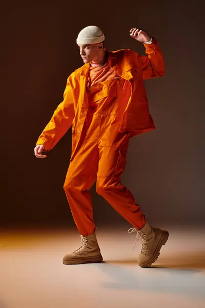 Молодой красивый мужчина в оранжевом комбинезоне и куртке, бежевая шапочка позирует на коричневом фоне — стоковое фото