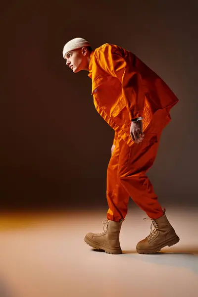 Vista lateral del hombre guapo en mono naranja y chaqueta, gorro beige de pie sobre fondo marrón - foto de stock