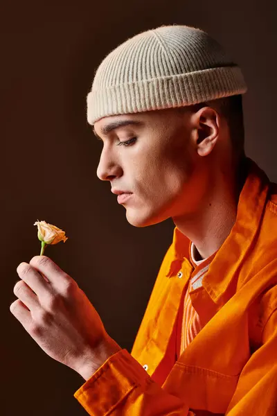 Vista lateral del hombre de moda en traje naranja con gorro beige sosteniendo la flor en la mano - foto de stock