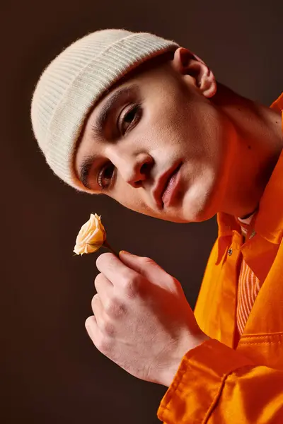 Handsome man in orange outfit wearing beige beanie holding flower in hand and looking at camera — Stock Photo