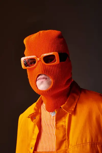 Elegante chico en traje naranja con mascarilla de pasamontañas y gafas de sol naranjas sobre fondo marrón - foto de stock