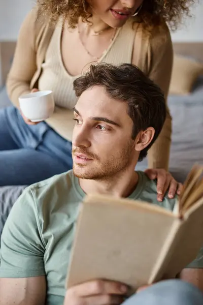 Relaxed brunette man holding book and looking away, hugged by his girlfriend, morning couple time — Stock Photo