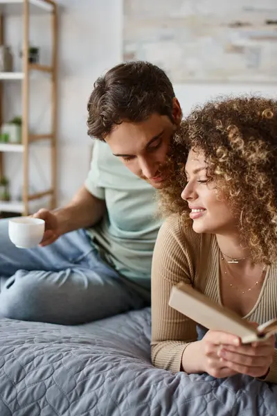 Cozy morning with curly young woman reading her book while being hugged by brunette man — Stock Photo