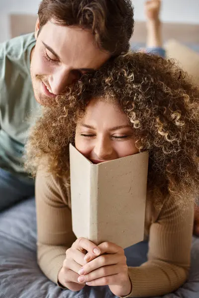 Dulce mañana abrazo de morena hombre como rizado joven mujer lee su libro en la cama - foto de stock